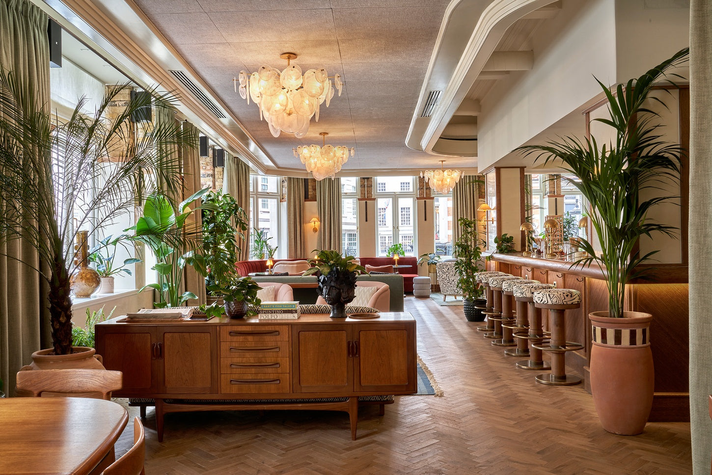A wide, elegant interior hallway with wood flooring, chandeliers, large potted plants, a wooden sideboard in the foreground, and a bar counter lined with patterned stools on the right.